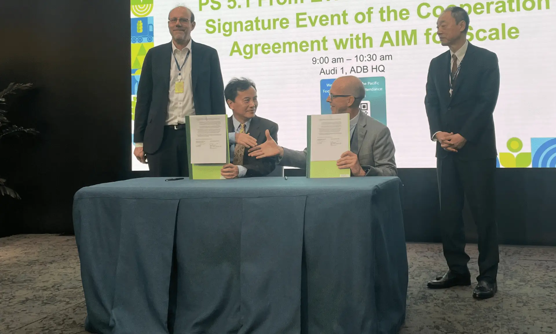 Two men behind desk shake hands and hold up documents. There are two men behind them on stage.