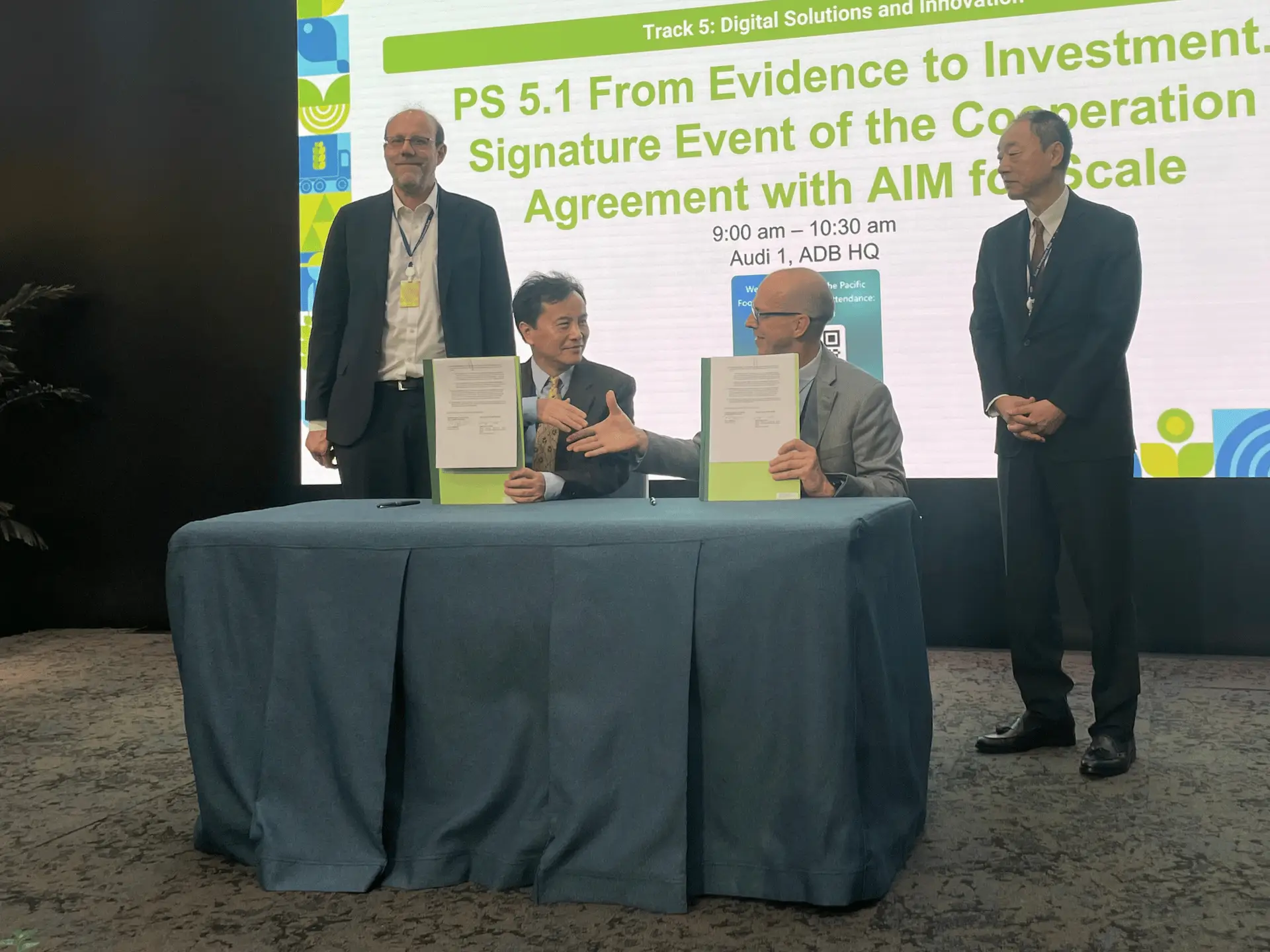 Two men behind desk shake hands and hold up documents. There are two men behind them on stage.